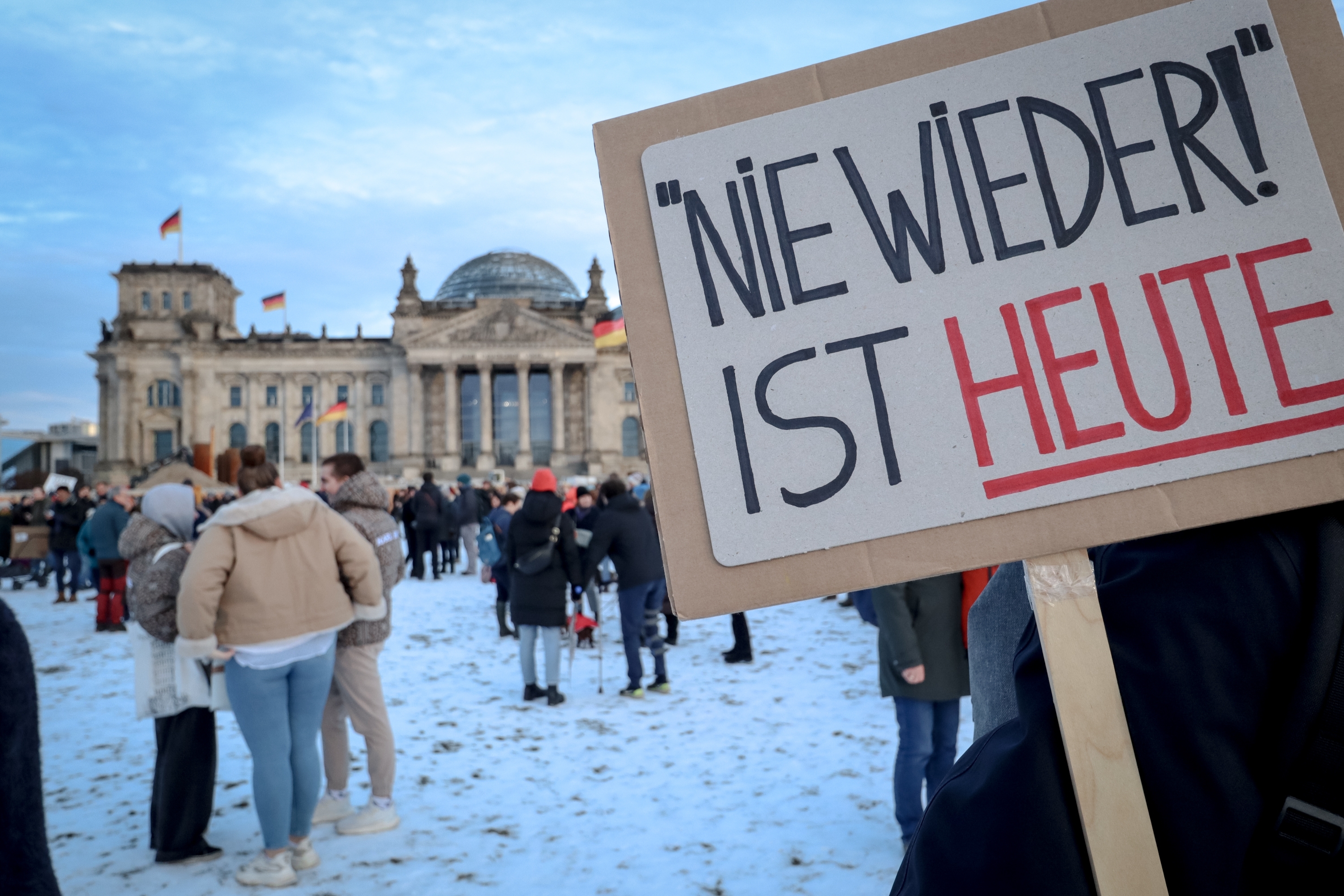 Demonstration vor dem Reichstagsgebäude in Berlin: Im Vordergrund ein Pappschild mit der Aufschrift „Nie wieder ist heute“, im Hintergrund Menschenmenge und deutsche Flaggen im Schnee.